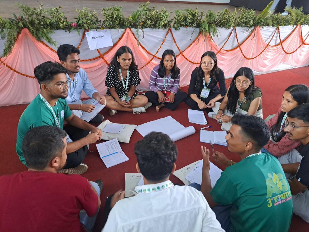 Young individuals sit on the ground in a circle with notebooks in front of them and a sign behind then reads 'Dhaulagiri', their group name. 